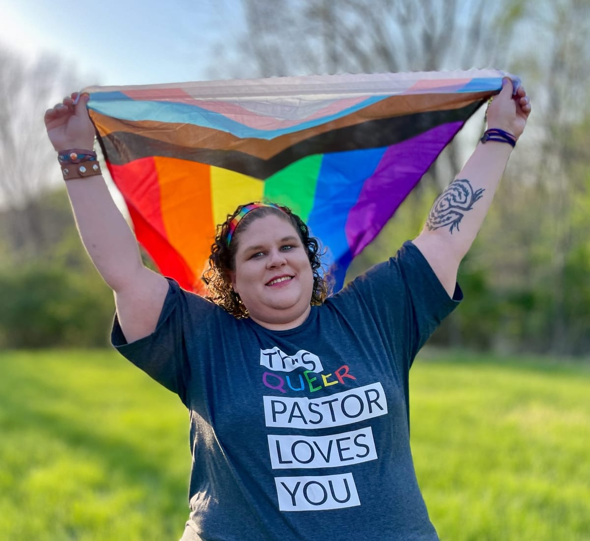 Sarah standing in a field in a shirt that says "THIS QUEER PASTOR LOVES YOU," holding a Progress LGBT flag over her head. A labyrinth tattoo is visible on her left arm. 